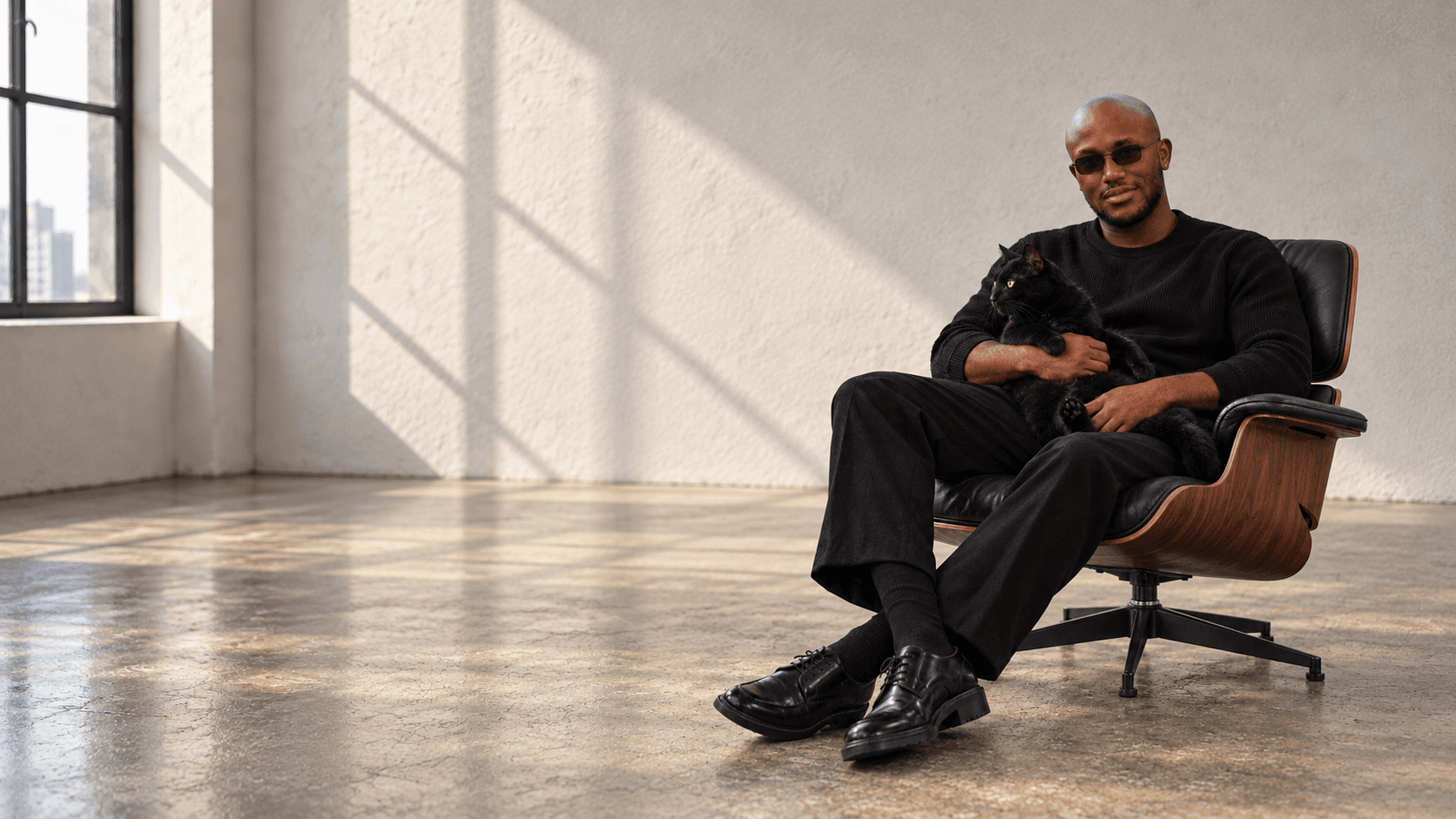 Muiz in black tailoring, seated with a black cat in a sunlit loft